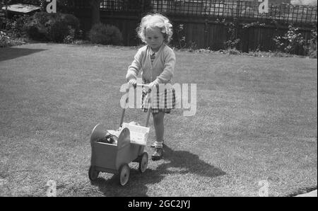 Anni '60, storico, fuori in un giardino posteriore sull'erba, una giovane ragazza che spinge il suo pram bambole in legno, Inghilterra, Regno Unito. Foto Stock