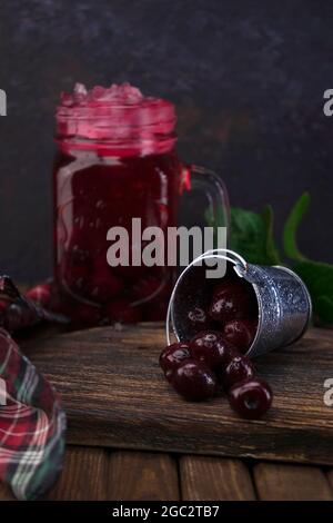 Un piccolo secchiello in alluminio con frutta fresca di ciliegia rossa organica sparsa su un vecchio pannello testurizzato di legno contro un vaso di vetro di cocktail di ciliegia con ghiaccio Foto Stock