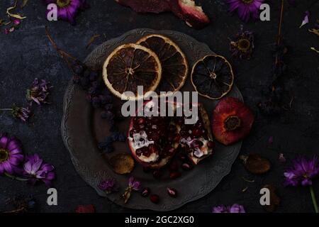 Melograno maturo in un piatto d'annata, un mazzo di uve secche e fette d'arancia tra fiori selvaggi su fondo di pietra scuro Foto Stock