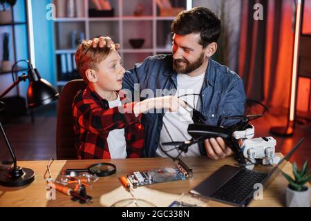 Uomo caucasico e ragazzino che riparano il quadcopter moderno mentre si siede a tavola. Piangendo padre insegnando il lavoro del figlio diverso dell'uomo a casa. Concetto parenting. Foto Stock