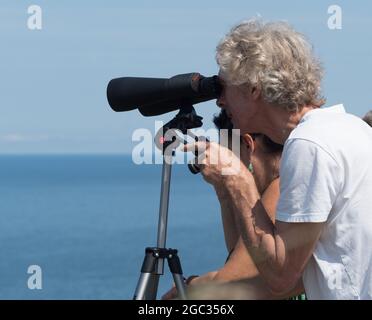 Un uomo guarda attraverso un grande paio di binocoli skymaster supportati su un Manfrotto tripod.Sea e oceano in backgroun Foto Stock