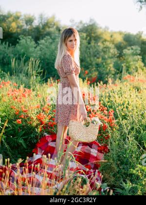 Donna bionda caucasica all'aperto in posa su plaid con fiori di papavero al tramonto. Donna in abito e borsa in vimini con fiori Foto Stock