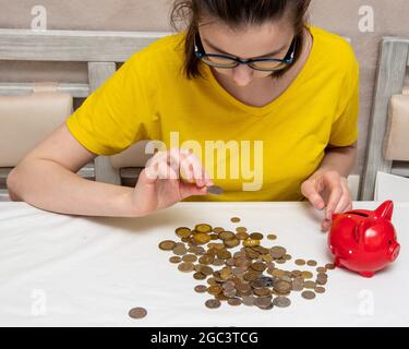 Ritratto di una giovane ragazza in una T-shirt gialla guarda una moneta, ci sono un sacco di monete sul tavolo, una banca piggy rosso. Foto Stock