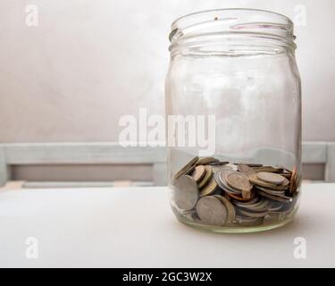 monete di diversi paesi in un classico vaso di vetro su sfondo grigio. Concetto di conservazione finanziaria. Foto Stock