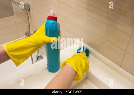 Le mani in guanti gialli puliscono il bagno bianco con un detergente e una spugna blu sull'impugnatura. Il concetto di lavoro a casa, disinfezione Foto Stock