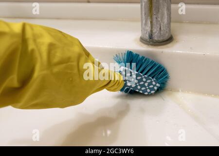 Una mano in un guanto giallo pulisce il lavandino con una spazzola blu sul manico, pulendo il bagno. Il concetto di lavoro a casa, disinfezione Foto Stock