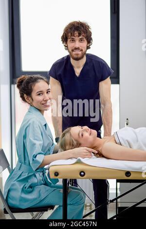 Due piacevoli massaggiatori praticano il massaggio sul viso e sulla testa del cliente femminile, il massaggiatore maschile e femminile in un lavoro uniforme insieme, la signora bionda giacente sul massag Foto Stock