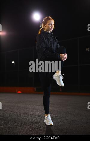 Giovane donna caucasica snella che fa l'allenamento stretching all'aperto nella strada della città di notte. Attraente donna focalizzata con capelli rossi naturali impegnati nello sport Foto Stock