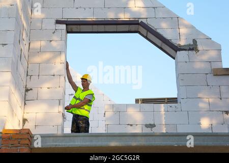 Il lavoratore di costruzione in cantiere misura la lunghezza dell'apertura della finestra e la parete con metro a nastro. Cottage sono fatti di cemento poroso Foto Stock