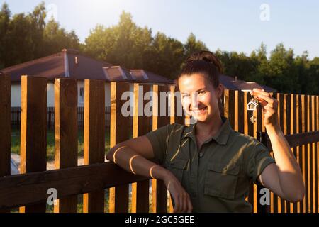 Donna felice con le chiavi della casa in mano vicino alla recinzione di legno della sua casa in un villaggio cottage. Proprietario, agente, immobiliare. Muovendosi, comprando un ho Foto Stock