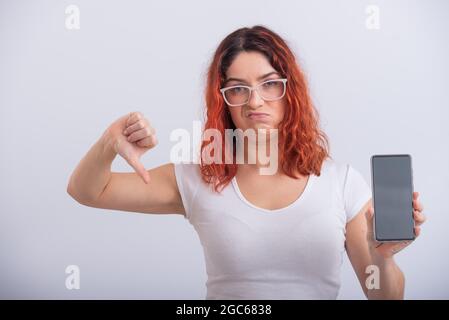 Donna caucasica che tiene uno smartphone con schermo nero e mostra il pollice verso il basso. Foto Stock