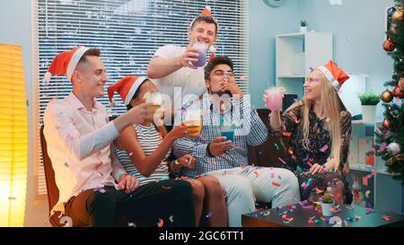 Primo piano di allegra azienda che celebra il Natale in confetti che soffiano mentre fanno gli cheers e soffiano fischietti di festa a casa. Foto Stock