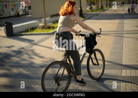 Una ragazza guida una bicicletta. Passeggiate per la città in bicicletta. Una ragazza con i capelli rossi sta guidando giù la strada. Foto Stock