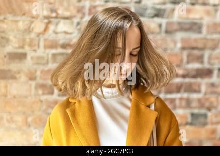 Ritratto di una giovane donna in un cappotto giallo luminoso contro lo sfondo di una parete di mattoni vicino alla finestra. Al sole Foto Stock