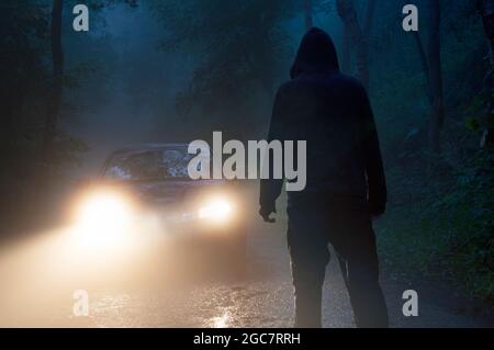 Una figura con cappuccio sinuosa che si contorna contro i fari di un'auto. Su una spaventosa strada forestale di notte. Foto Stock