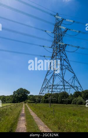 Un sentiero in campagna che corre sotto linee elettriche ad alta tensione che si estendono attraverso un campo di fattoria al sole estivo. Foto Stock