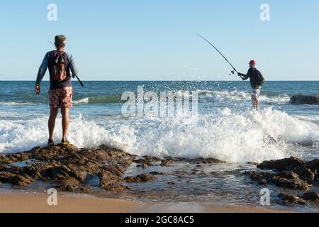 Salvador, Bahia, Brasile - 25 aprile 2021: Pescatori su rocce con i loro pali di pesca cercando di catturare il pesce da vendere e consumare. Foto Stock