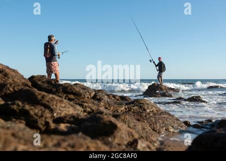 Salvador, Bahia, Brasile - 25 aprile 2021: Pescatori su rocce con i loro pali di pesca cercando di catturare il pesce da vendere e consumare. Foto Stock