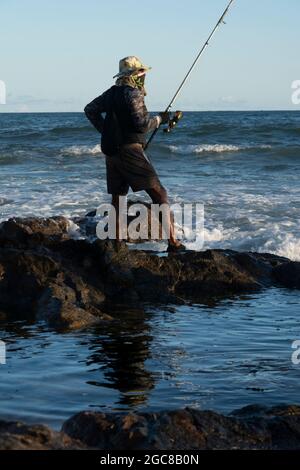 Salvador, Bahia, Brasile - 25 aprile 2021: Pescatori su rocce con i loro pali di pesca cercando di catturare il pesce da vendere e consumare. Foto Stock