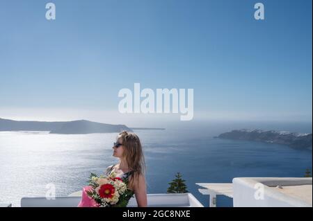 Giovane donna in occhiali da sole con bouquet di bellissimi fiori sulla terrazza del lussuoso hotel sull'isola di Santorini. Vista sulla caldera. Mar Egeo. Foto Stock