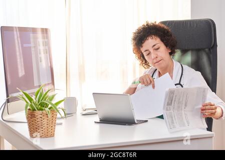 Donna medico digitalizzare i documenti attraverso il suo tablet seduto nel suo ufficio Foto Stock