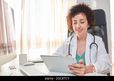 Una dottoressa usa il suo tablet digitale seduto alla scrivania del suo ufficio medico Foto Stock