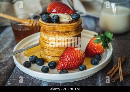 Vista in primo piano su una pila di pancake con bastone di miele, fragola, bacca blu e panna a frusta in cima al piatto, delizioso dessert con latte per colazione. Foto Stock
