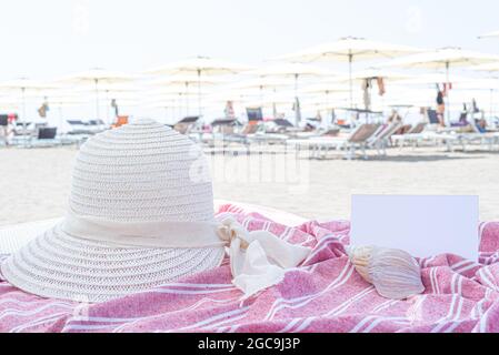 Vacanza estiva ancora vita. Cappello di paglia bianca, conchiglia su tovagliolo rosso e bianco.ombrelloni da spiaggia sullo sfondo. Biglietto di invito o biglietto di auguri vuoto Foto Stock