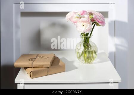 Un bouquet di bicchierini rosa persiani in un vaso di vetro e due regali avvolti su un tavolo bianco di fronte ad un falso camino bianco Foto Stock