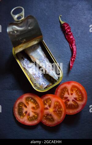 Primo piano di una lattina di sardine accanto ad alcuni pomodori rossi e un peperoncino su un'ardesia nera Foto Stock