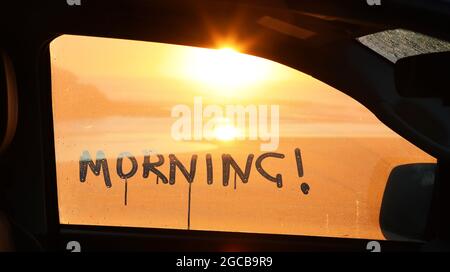 La parola mano mattina scritta nella condensazione fuori di una finestra di auto con una bella alba dorata mattina sulla costa vista fuori dalla finestra. Foto Stock