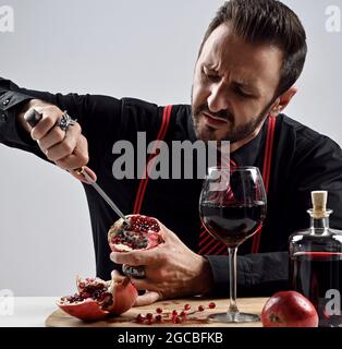Brutale uomo bearded in camicia nera con cravatta a righe e sospetti al tavolo mangia fresco bastoni di melograno coltello in esso Foto Stock