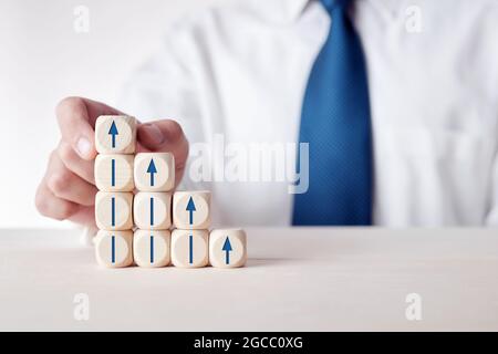 Uomo d'affari che mette il cubo con l'icona della freccia sulla parte superiore dei cubetti di legno a più livelli. Concetto di crescita economica, finanziaria o professionale, sviluppo o progresso. Foto Stock