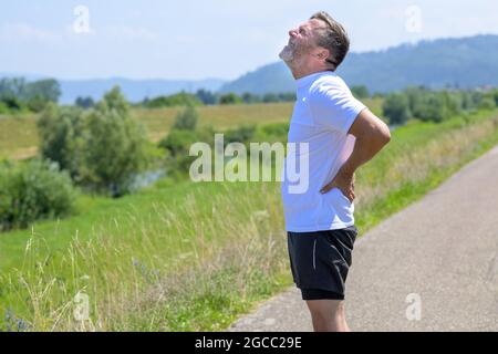 Uomo di mezza età che soffre di dolore acuto alla schiena che gli stringe la parte inferiore della schiena con le mani e che si lamenta mentre si riposa dallo scherzo quotidiano nel paese Foto Stock