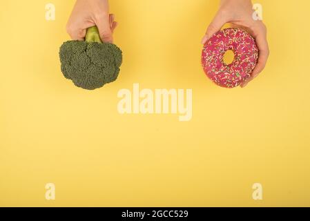 Confronto delle abitudini alimentari. Donna che tiene broccoli e ciambella su sfondo giallo. Spazio di cimasa. Foto Stock