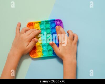 Le mani dei bambini giocano il giocattolo popit, vista dall'alto su sfondo blu a due ombre. Popit su sfondo blu, spazio di copia. Popolare giocattolo in silicone colorato antistress pop it per bambini. Vista dall'alto. Concavità semplice Foto Stock