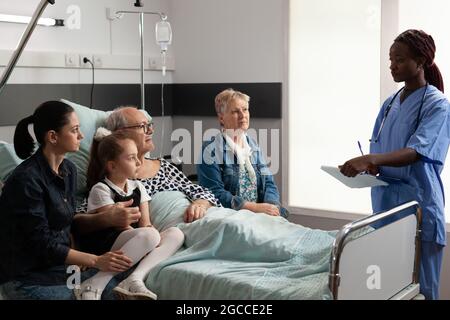 Infermiera afroamericana che discute del trattamento di malattia con l'anziano anziano anziano nel reparto dell'ospedale. Una famiglia allegra e premurosa, seduta accanto a un paziente malato, lo sostiene durante il recupero clinico Foto Stock