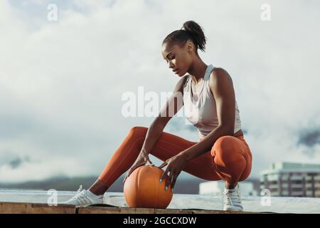 Donna sportiva con pallacanestro seduta sul tetto. donna in forma fisica che si rilassa dopo una sessione di allenamento all'aperto. Foto Stock