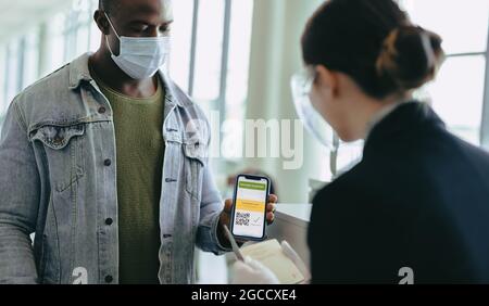 Uomo con maschera facciale che tiene il telefono cellulare per mostrare il passaporto del vaccino al personale dell'aeroporto al banco del check-in. Operatore di terra che visualizza il passaporto del vaccino di ma Foto Stock
