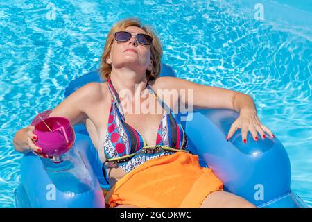 Una donna bianca di mezza età si rilassa sdraiata su un materasso gonfiabile sull'acqua di una piscina, con un bicchiere di cocktail in mano Foto Stock