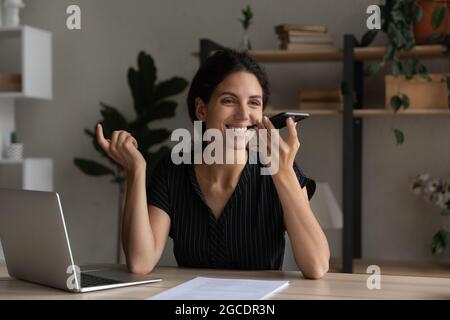 Donna d'affari sorridente che registra messaggi vocali sullo smartphone, seduto alla scrivania Foto Stock