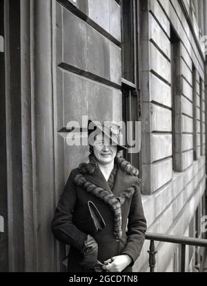 Anni '30, storica ed elegante donna che indossa un cappello con una piuma e vestito in abito, con nappine decorative in pelliccia, in piedi presso un ingresso in una terrazza georgiana, Scozia, Regno Unito Foto Stock