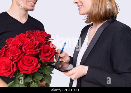 L'uomo sorridente del corriere ha trasportato un bouquet bello delle rose rosse ad una donna bionda e deve mettere la sua firma sul tablet per il servizio. Foto Stock