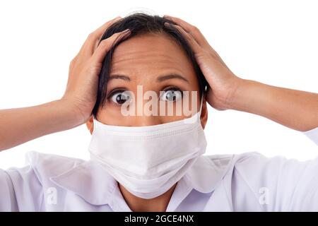 Donna medico indossando coronavirus maschera chirurgica viso in panico paura di fare una stressata espressione facciale tenendo la testa da mal di testa. Foto Stock