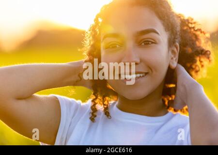 Bella biraciale misto razza Africano Americano adolescente ragazza giovane donna sorridente fuori retroilluminata al tramonto o all'alba Foto Stock