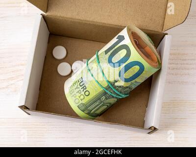 Rotolo di banconote da 100 euro e poche pillole in una piccola scatola di cartone aperta. Risparmio di denaro per l'assistenza sanitaria e il trattamento, la pianificazione per il concetto di spese. Foto Stock