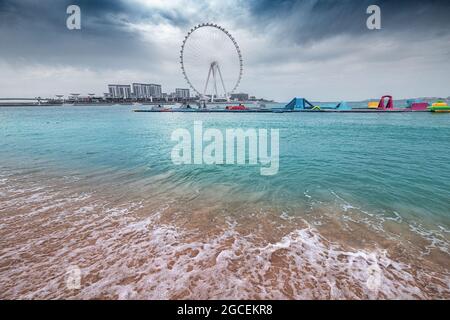 Un'onda si schianta sulla spiaggia di sabbia e sulla famosa ruota panoramica del Dubai Eye durante il tempo nuvoloso Foto Stock
