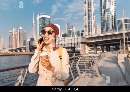 Asian Woman in un headscarf bere caffè e parla e gossip al telefono con un amico sul canale Dubai Walk Foto Stock