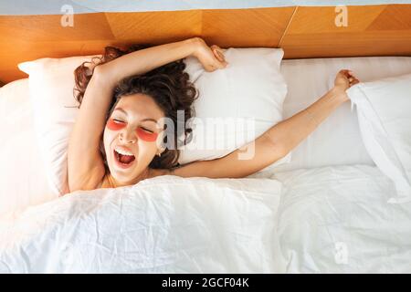 donna felice con le macchie degli occhi sbadiglia e si allunga mentre incontra la mattina nel suo letto. Concetto di sonno e sonnellino Foto Stock