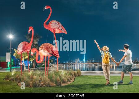 Felice coppia turistica passeggiate nella zona di Dubai Creek Marina Harbour e si pone sullo sfondo di giganteschi fenicotteri e la Torre Burj Khalifa Foto Stock